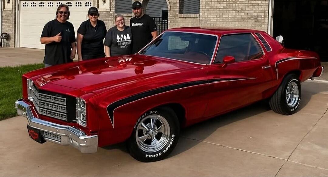 Four people, two men and two women, are standing next to a custom red classic car with black stripes. The car has chrome details and 'COBRA' written on its tires. The people are smiling, and one man is giving a thumbs-up.