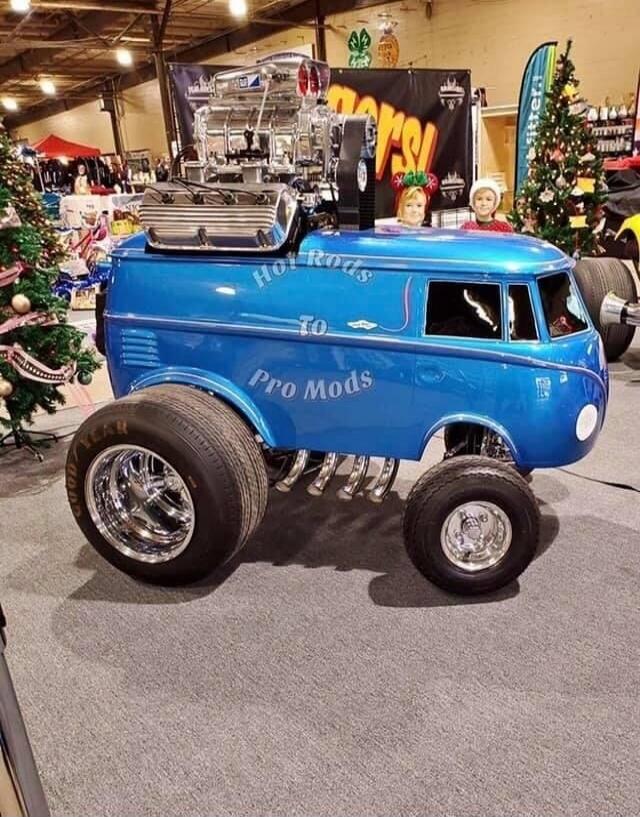 Hot Rods To Pro Mods. A highly customized blue van, resembling a modified Volkswagen bus, with a large exposed engine, big rear wheels, and smaller front wheels. Two children are visible in the front windows, wearing festive headwear. The scene appears to be an indoor car show or exhibition with Christmas trees in the background.