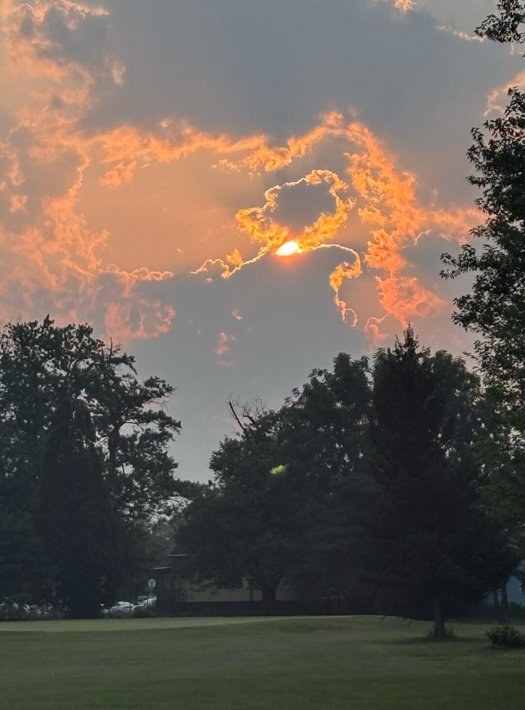 Sunset sky with orange clouds over a grassy field and trees.