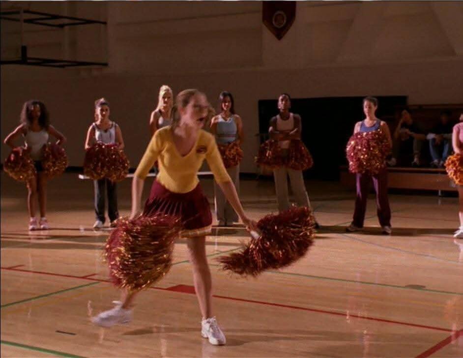 The cheerleaders are practicing their routine in a gymnasium. They are all wearing similar uniforms and holding pom-poms. The main cheerleader in the foreground is in motion, while the others are standing behind her.