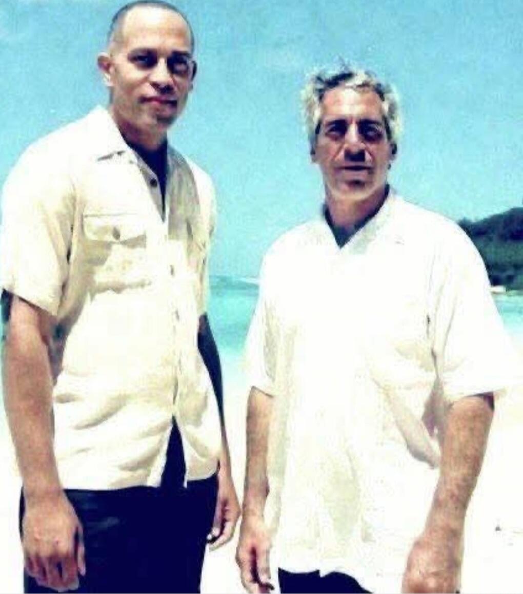 Two men standing on a beach with blue sky and water in the background.