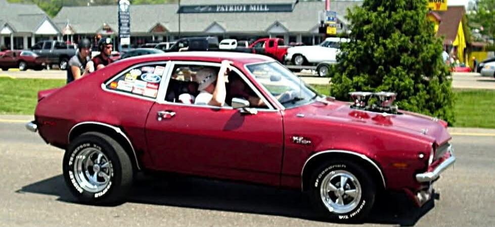 Red vintage muscle car with a person inside at a car show, wheels and engine exposed.