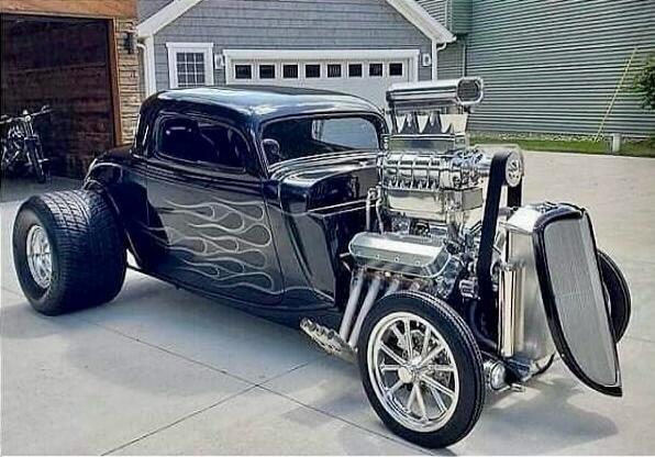 A custom black hot rod with a large chrome engine and flame decals is parked on a driveway in front of a garage.