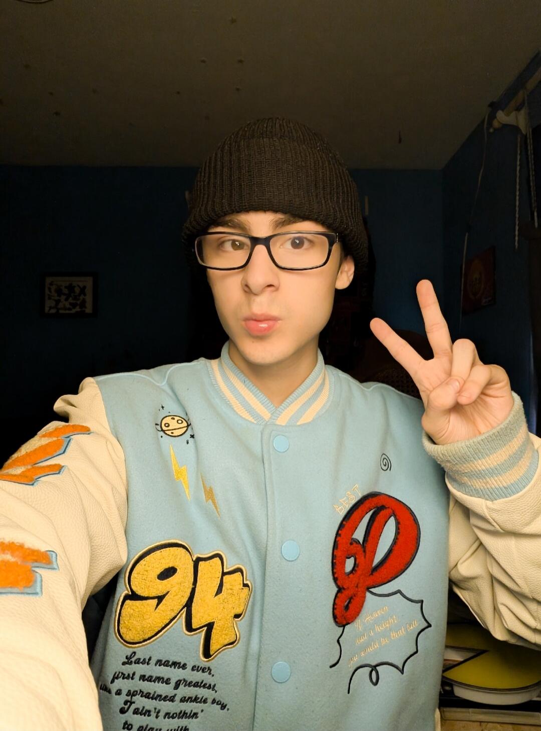 A young person wearing a black beanie, glasses, and a light blue and cream varsity jacket. They are making a peace sign with one hand and pouting their lips. The jacket features patches and embroidered text including '94', 'Last name ever, first name greatest, a sprained ankle boy, I ain't nothin' to play with', 'BEST', and 'If Heaven was a height,