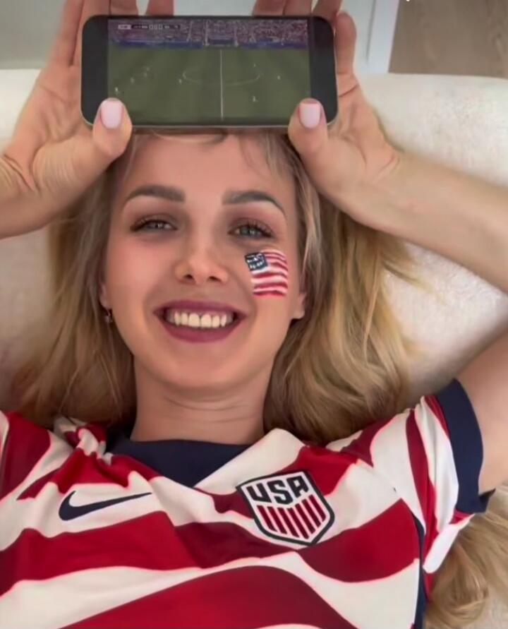 A woman is wearing a USA jersey and has an American flag painted on her cheek. She is watching a soccer match on a smartphone.