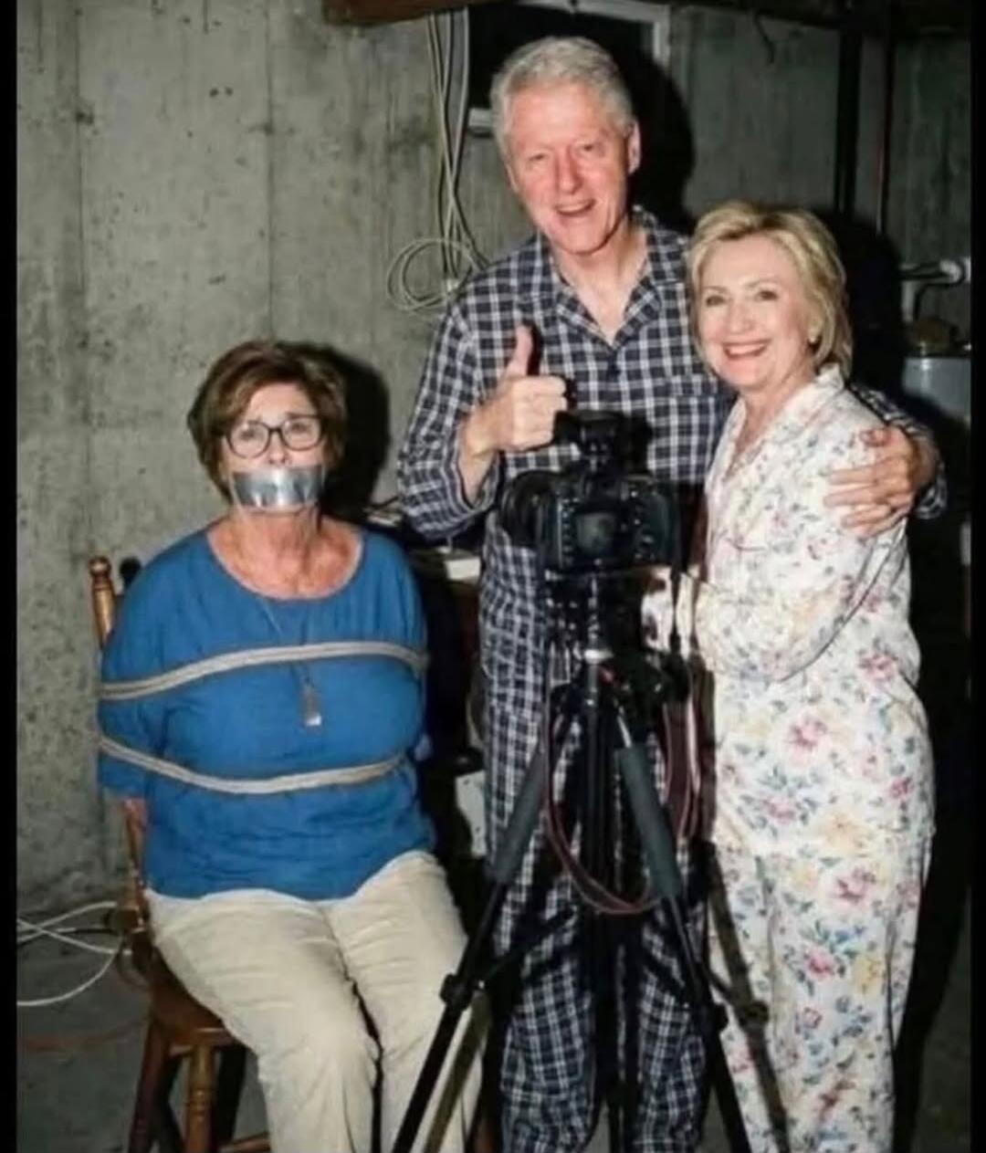 Three people in a basement setting; a woman sits on a chair with duct tape over her mouth, a man stands behind a camera giving a thumbs up, and another woman stands beside him smiling.