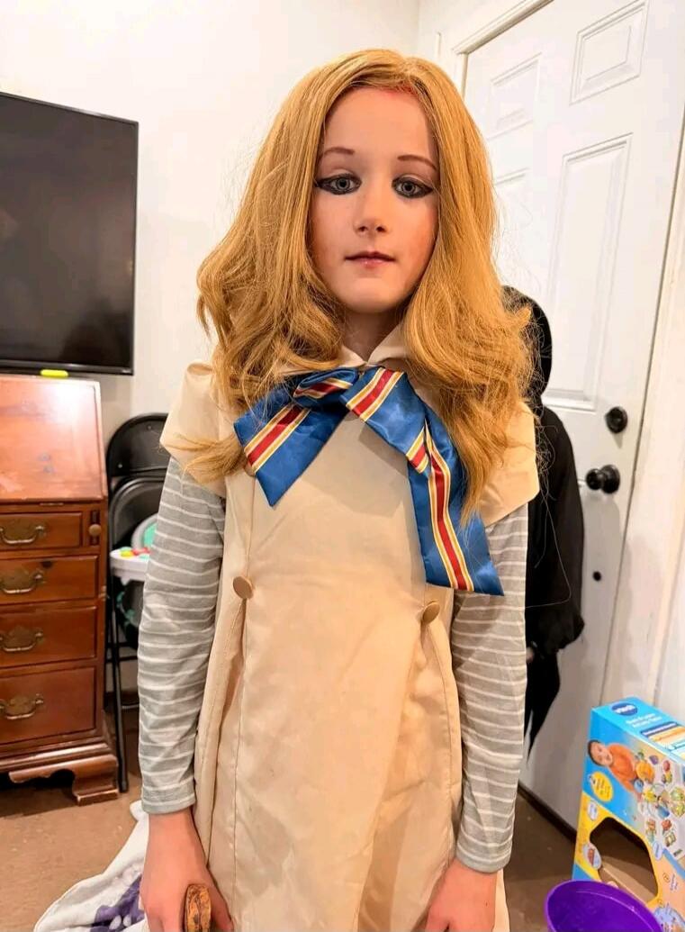 A child dressed in a M3GAN-like costume, featuring a blonde wig, heavy eye makeup, a beige dress with a blue and red bow, and a striped long-sleeve shirt underneath. The child is standing indoors in what appears to be a living room or bedroom, with a TV, dresser, and white door visible in the background.