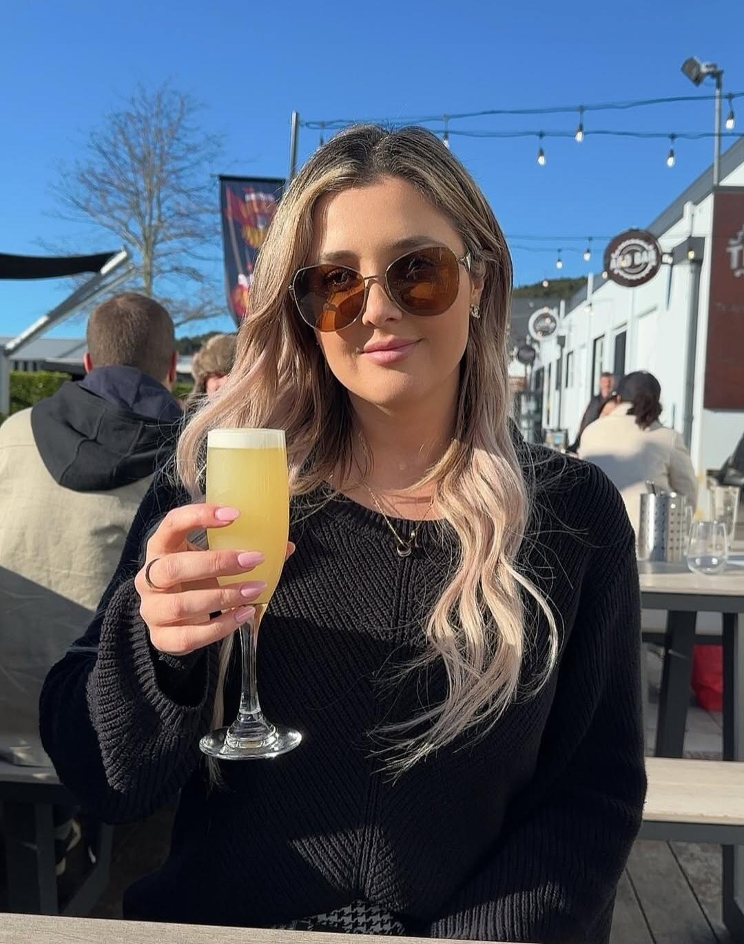 A woman wearing sunglasses and a black sweater is sitting outdoors, holding a glass with a yellow drink. The background shows an outdoor seating area with other people and buildings under a clear blue sky.