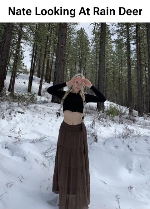 Nate Looking At Rain Deer. A young woman with blonde braids, wearing a black crop top and a long brown skirt, stands in a snowy forest, making a heart gesture with her hands.