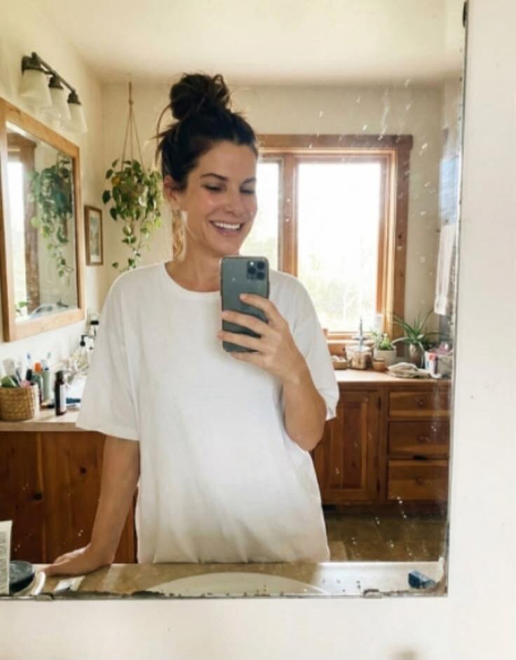 A woman is taking a mirror selfie in a bathroom. She is smiling and wearing a white t-shirt, with her hair tied in a bun. The background shows a wooden vanity, plants, and a window.