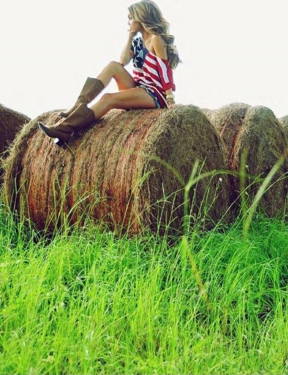 A person wearing a shirt with the American flag pattern sits on top of a large hay bale in a grassy field, wearing brown cowboy boots.