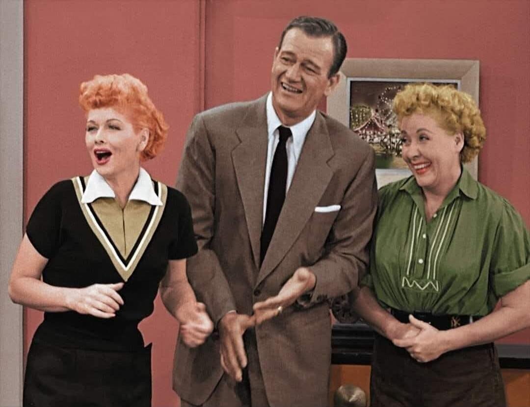 Three classic TV stars, Lucille Ball, John Wayne, and Vivian Vance, are laughing and smiling together. Lucille Ball is on the left, wearing a black and gold V-neck top. John Wayne is in the center, wearing a suit and tie, with a wide smile. Vivian Vance is on the right, wearing a green shirt, also smiling broadly. They appear to be enjoying a momen