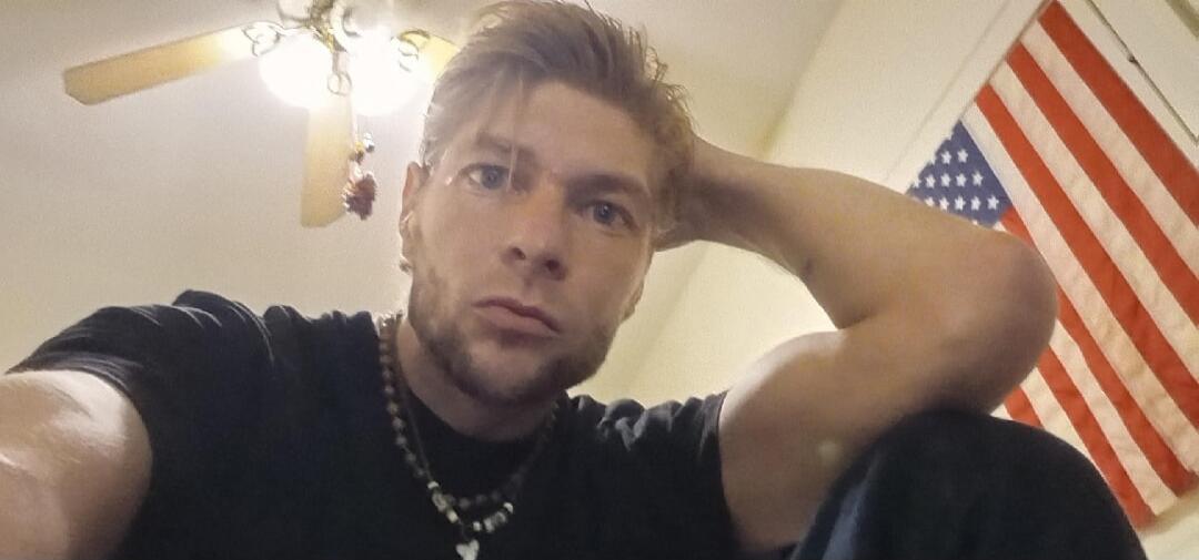 A man takes a selfie from a low angle, looking directly at the camera. He has light hair and a beard, wearing a black t-shirt and a beaded necklace. An American flag is visible in the background.
