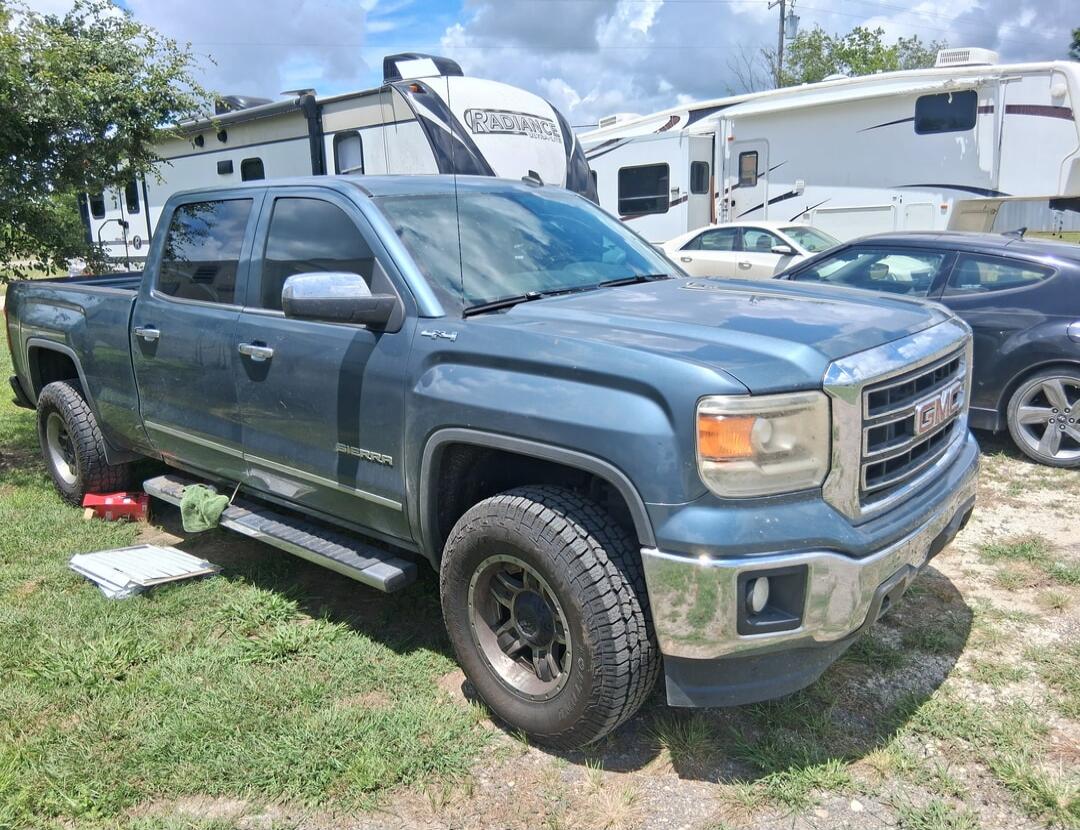 A blue GMC Sierra 4x4 pickup truck is parked on a grassy area. In the background, there are several recreational vehicles (RVs), one of which has 'Radiance' written on it. Other cars are also visible. There are red blocks under the rear tire of the truck, and a green cloth and some metal parts are on the ground nearby.