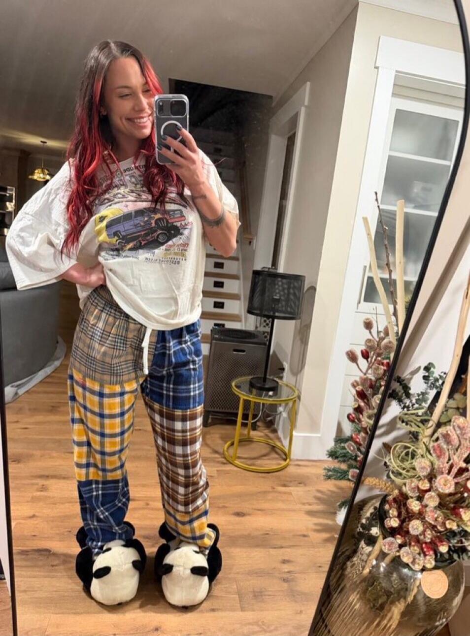 A woman with red highlights in her hair takes a mirror selfie, smiling. She is wearing an oversized white t-shirt with a graphic print, patchwork plaid pajama pants, and fluffy panda slippers. The background shows a cozy home interior.
