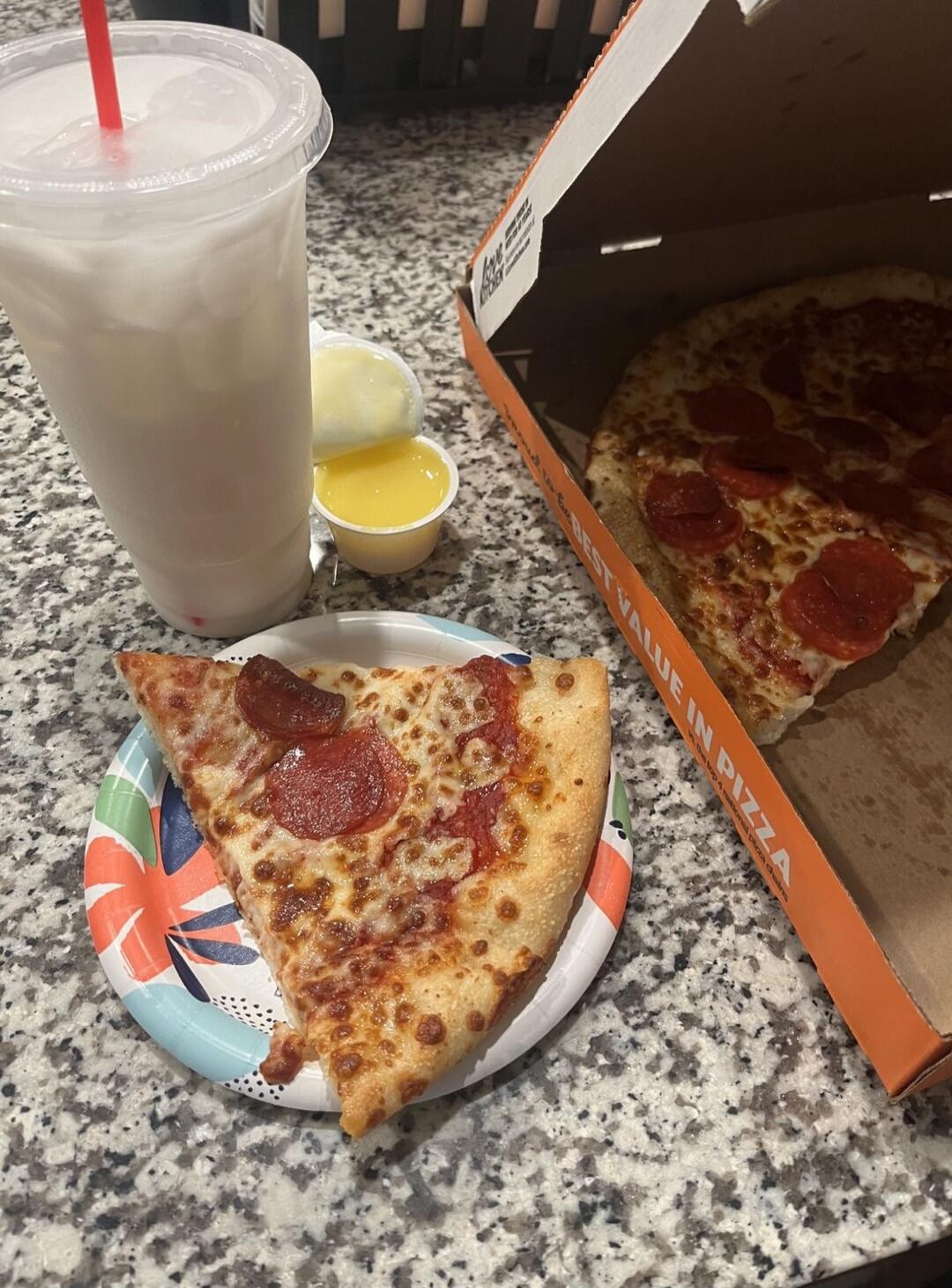 A meal consisting of a slice of pepperoni pizza on a colorful paper plate, a pizza box containing a whole pepperoni pizza, a large plastic cup with a drink and a red straw, and two small containers of dipping sauce. The text 'BEST VALUE IN PIZZA' is visible on the side of the pizza box.