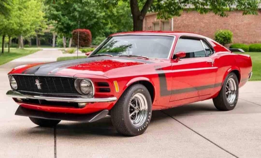 A classic red Ford Mustang Boss 302 with black racing stripes is parked on a paved driveway.