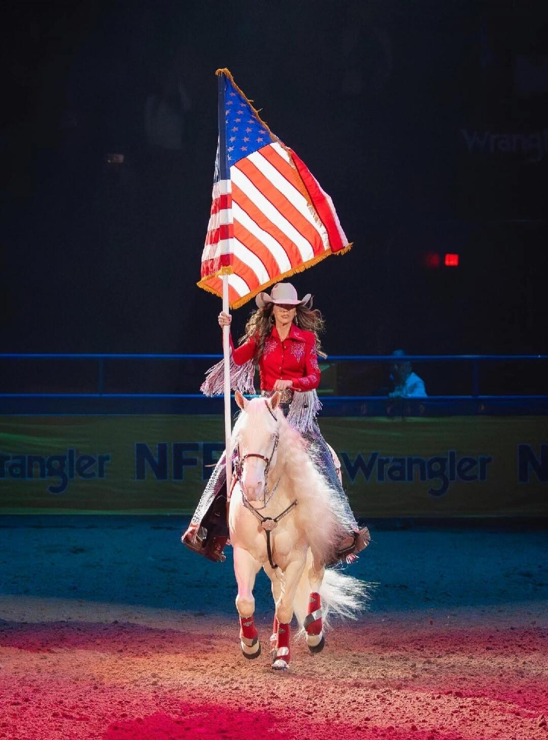 A woman in a cowboy hat and a red shirt rides a white horse while holding a large American flag. The background shows 'Wrangler NFR'.
