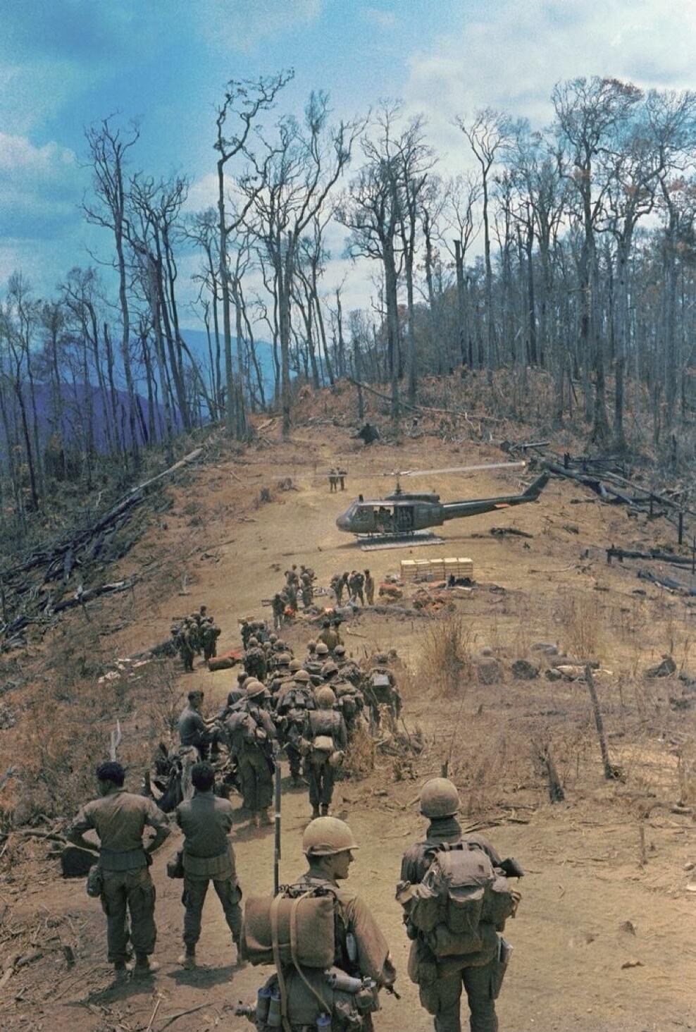 A group of soldiers are walking down a dirt path towards a helicopter. The helicopter is on the ground and appears to be waiting for the soldiers. The soldiers are carrying backpacks and are wearing camouflage uniforms. The landscape is dry and barren, with dead trees in the background. The sky is blue with some clouds.