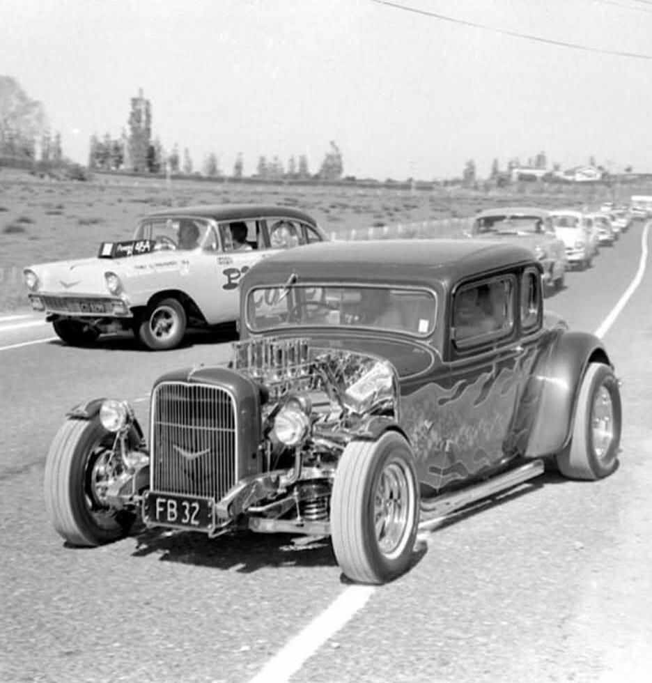 FB 32, 46A, P, COX. Two classic cars, a hot rod with an exposed engine and flame paint job in the foreground, and a 1957 Chevrolet in the background. They are on a road, with a line of other vintage cars in the distance. The hot rod has a prominent grille and large tires.