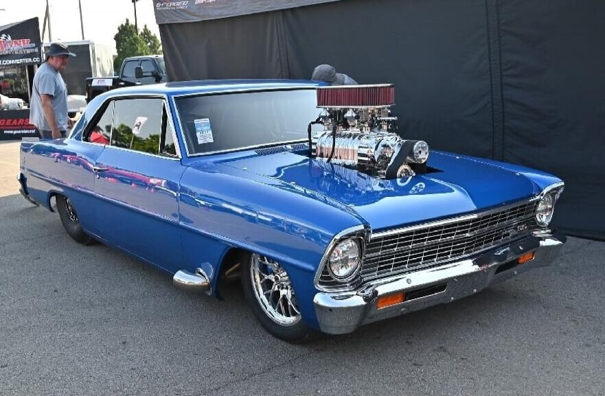 A blue classic car, possibly a Chevrolet Nova, with a large supercharger engine protruding from the hood. A man in a hat is standing near the car. In the background, there are banners with text like 'ENFORGED' and 'GEARS'.
