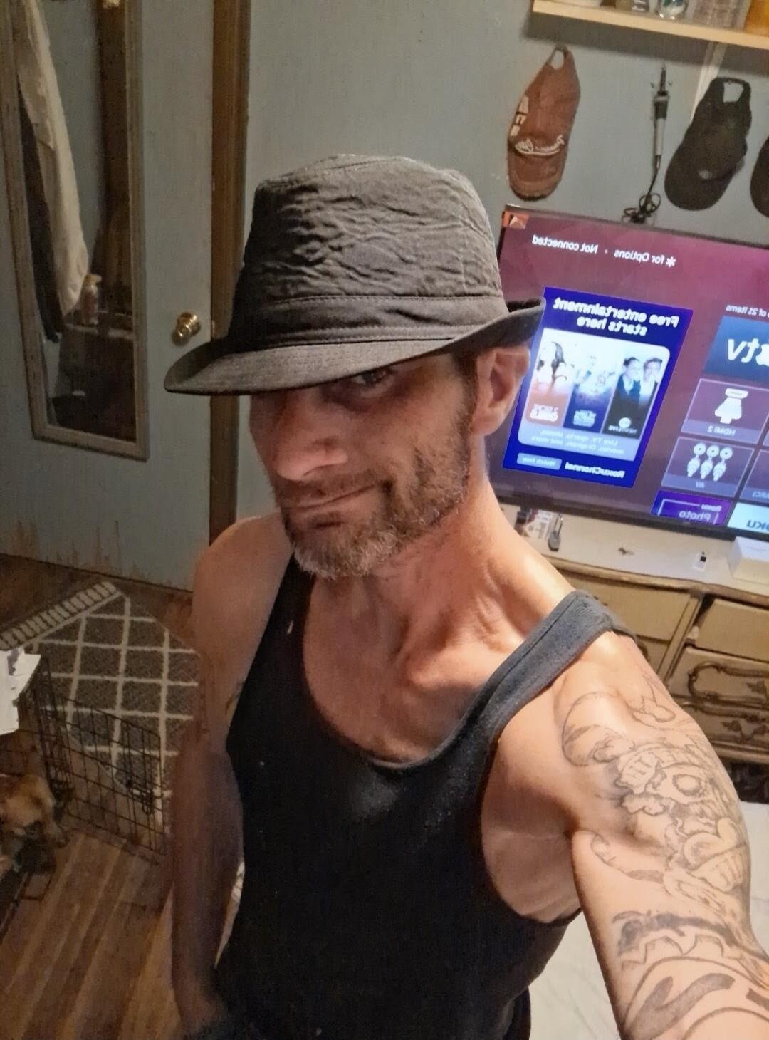 A man wearing a dark hat and a black tank top, with a beard and tattoos on his left arm, is taking a selfie. He is looking directly at the camera with a slight smile. In the background, a TV screen shows mirrored text, possibly 'Entertainment' and 'Options'. A mirror and some household items are also visible.