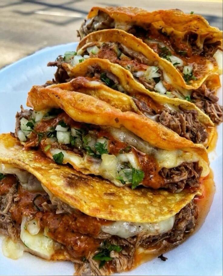 A stacked plate of crispy tortillas filled with shredded beef, melted cheese, onions, cilantro, and salsa. The stack resembles multiple tacos layered on top of each other.