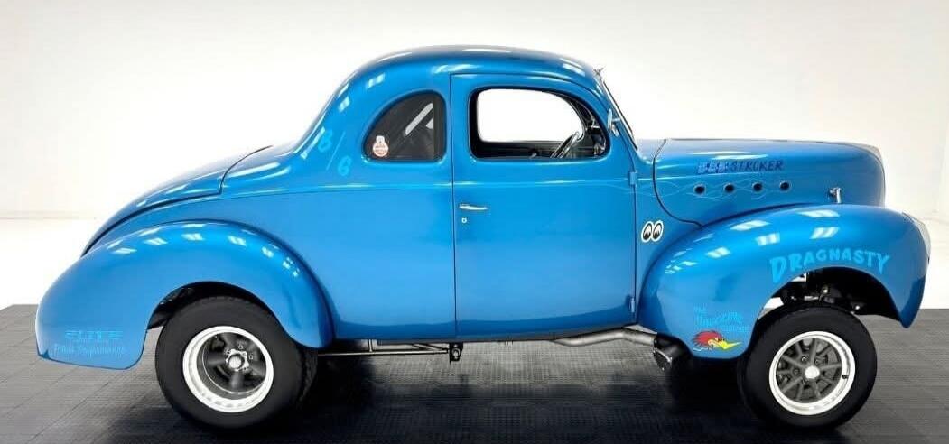 SELF-STROKER DRAGNASTY ELITE 6. A bright blue vintage-style drag racing car, possibly a gasser or hot rod, with custom lettering and a Road Runner cartoon character sticker.