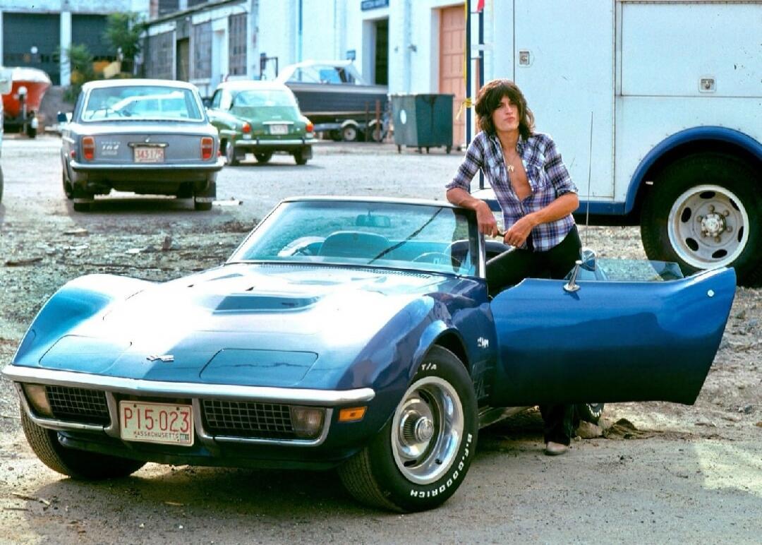 A person with long, shaggy hair and an unbuttoned plaid shirt is leaning on a blue vintage Chevrolet Corvette convertible with the driver's door open. The car has a Massachusetts license plate that reads 'P15-023'. Other vehicles and buildings are visible in the background.