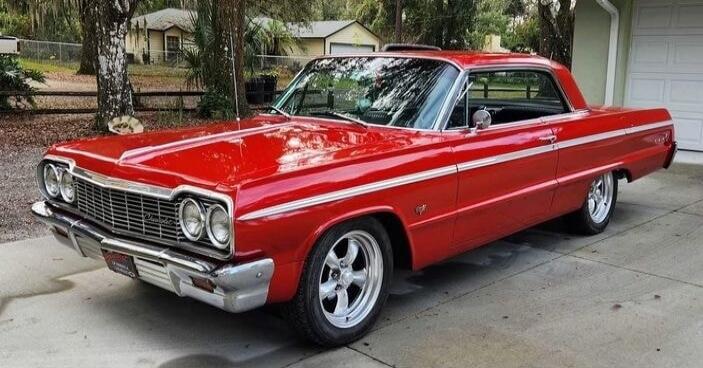 Red vintage car with chrome bumper parked in a suburban driveway.