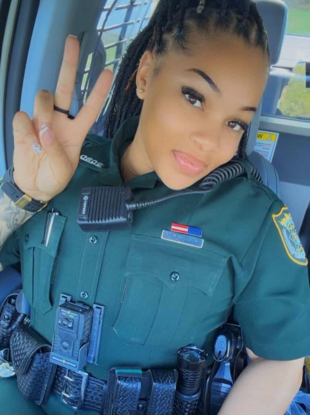 A woman in a police uniform making a peace sign. Text visible on uniform includes 'OSPD' and 'NEWMAN'. A body camera with 'ORALID HOTIAN' is attached to her belt. A warning sign with 'W A' is in the background.