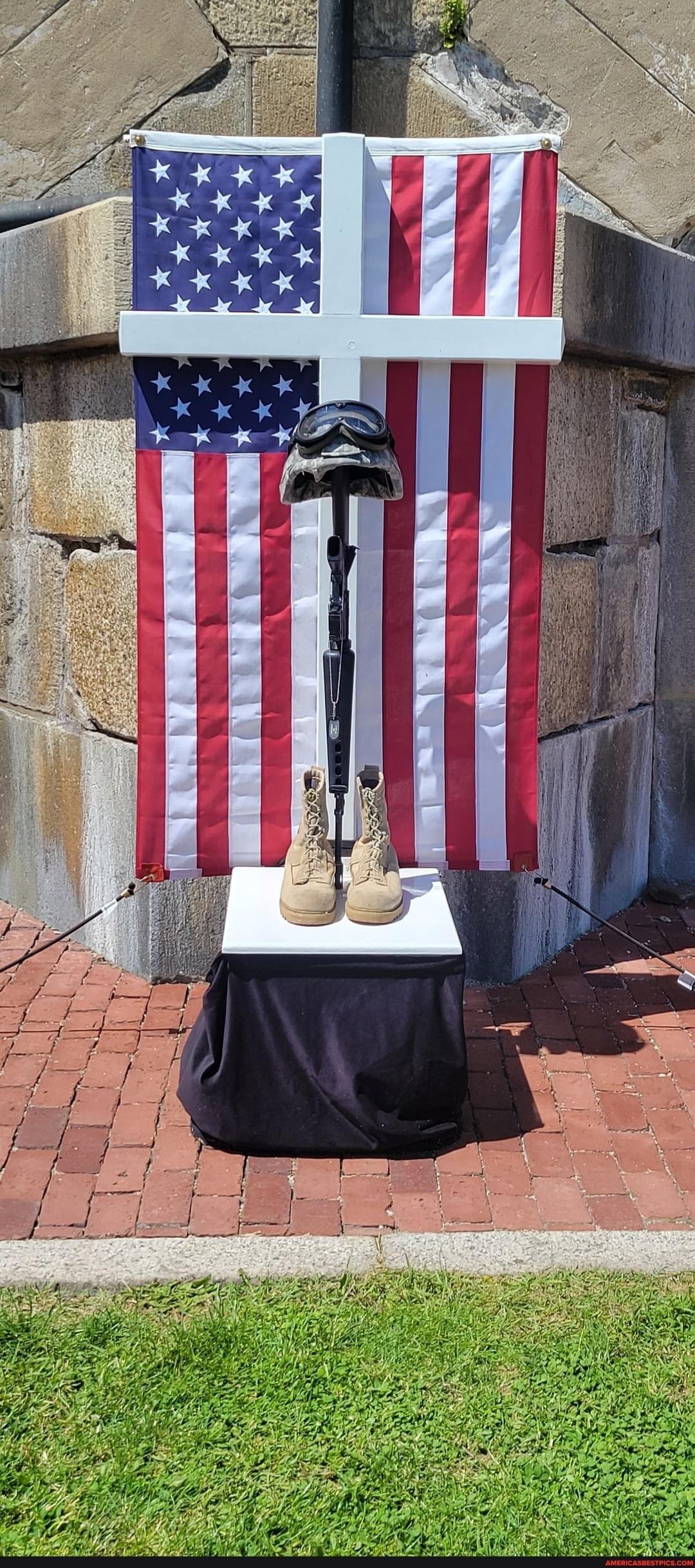 American flag backdrop with a white cross, a prosthetic leg, helmet, and boots on a pedestal.