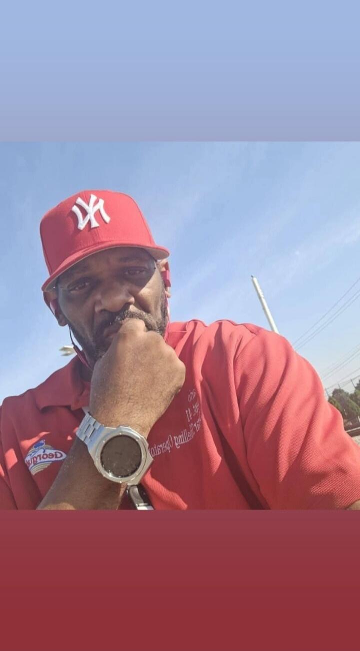 A man wearing a red New York Yankees baseball cap and a red polo shirt with a 'Geogrid' logo (reversed) on it. He has a beard and a silver watch on his wrist. His fist is near his chin, and he is looking at the camera under a clear blue sky.