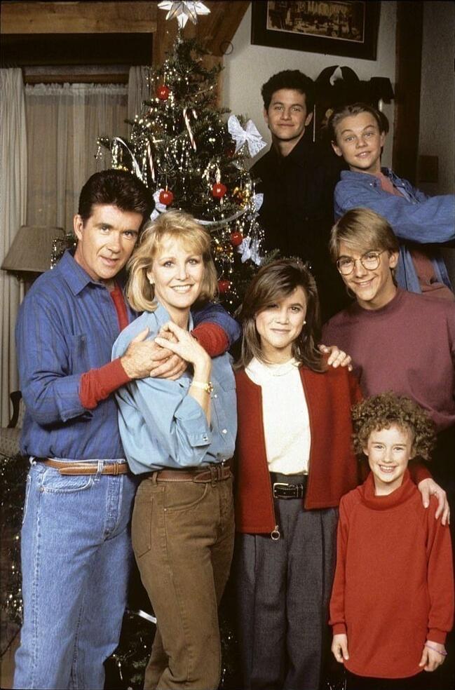 A family photo around a decorated Christmas tree in a cozy living room. Several adults and children stand together smiling, with festive attire and a warm holiday atmosphere.