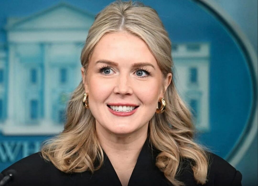 A smiling blonde woman with gold hoop earrings sits at a podium in front of a blue backdrop.