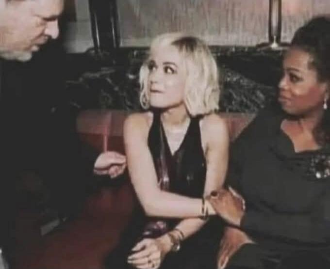 A man, widely identified as Harvey Weinstein, leans over a blonde woman, believed to be Rita Ora, who appears uncomfortable. Another woman, identified as Oprah Winfrey, holds Rita Ora's hand, seemingly offering support or comfort.