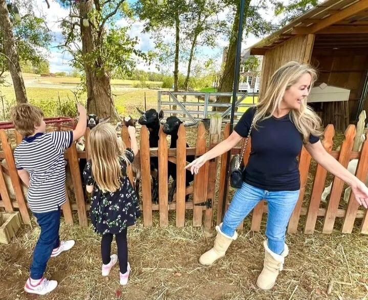 A woman in a black shirt and jeans stands near a wooden fence with children feeding or petting goats in a farmyard setting. There are trees and a small barn in the background.
