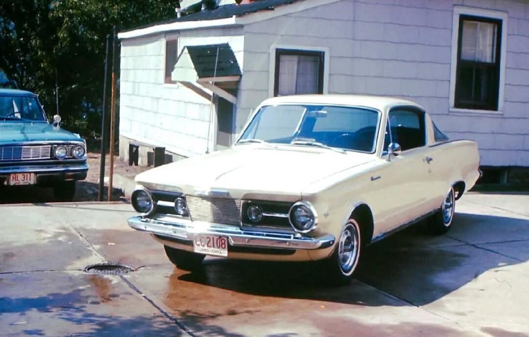 White vintage car parked on a driveway in front of a small house, with another blue car partially visible on the left.