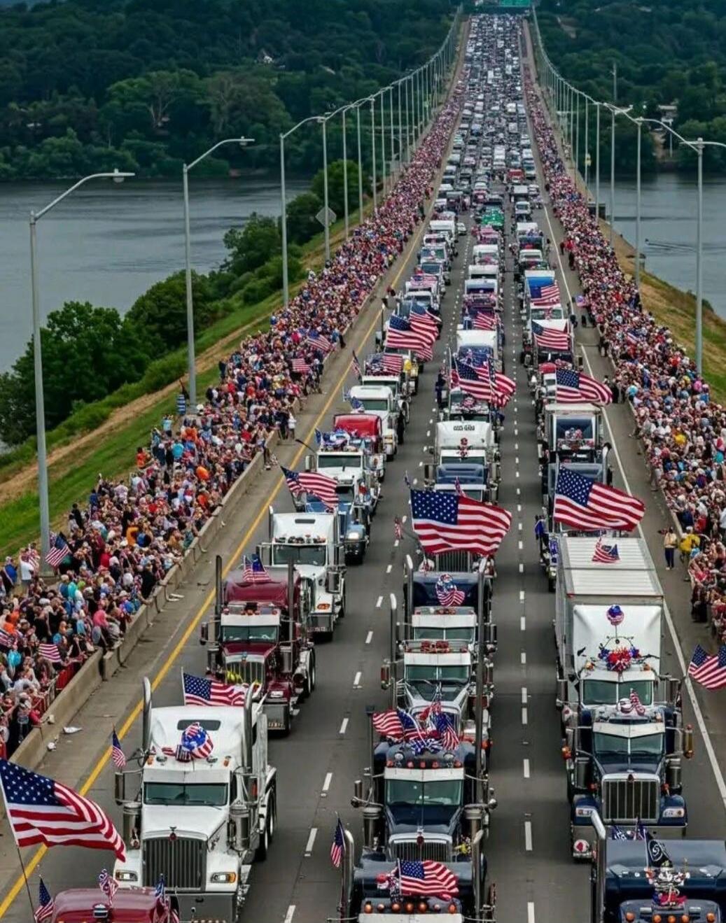 A long procession of trucks with American flags on a bridge, flanked by large crowds on both sides; a river runs beneath the bridge.