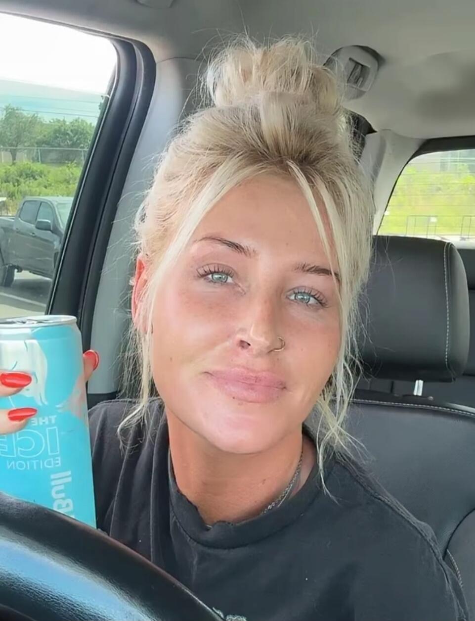 A blonde woman in a car holding a light blue can that says 'ICE EDITION' and 'lluB' (likely 'Bull' reversed). She has her hair in a bun, a nose piercing, and red nail polish.