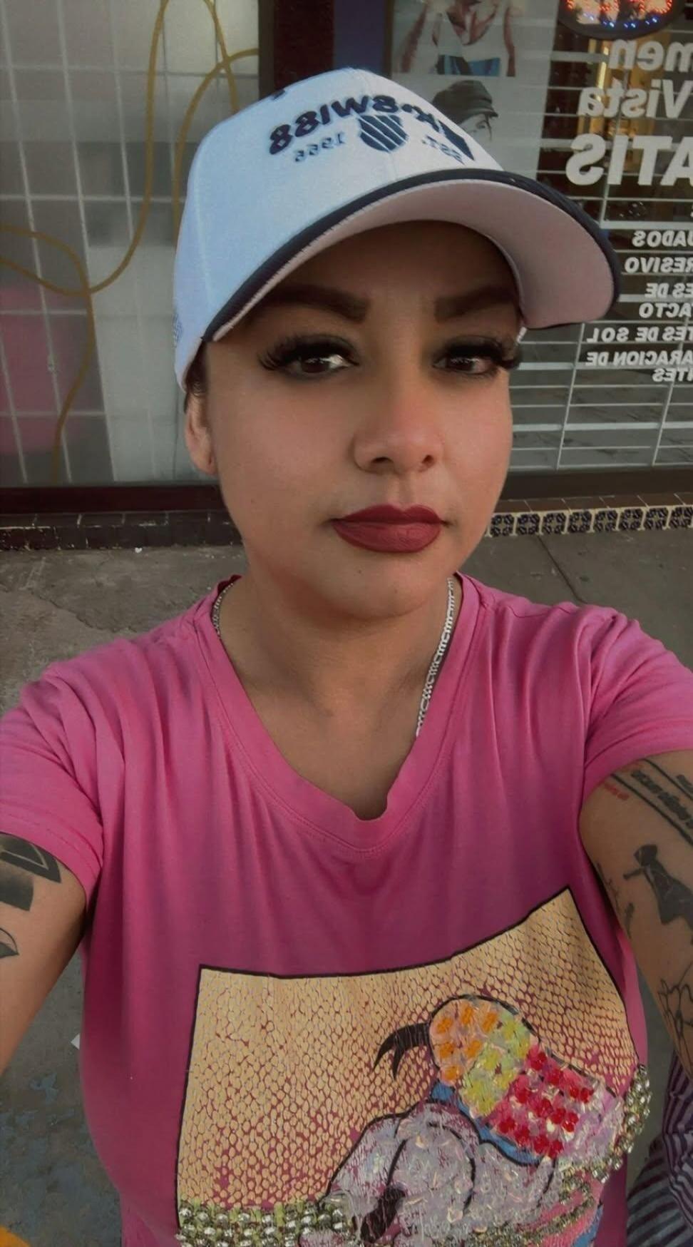 A woman wearing a white cap with 'SWISS 1988' written on it (reversed in the image), a pink t-shirt with a sequined design, and a silver chain. She has dark red lipstick and long eyelashes. Tattoos are visible on her arms. In the background, there's a reflection of text in a window, including words like 'VISITAS', 'GUADALUPE', 'SERVICIO', 'DE 201',