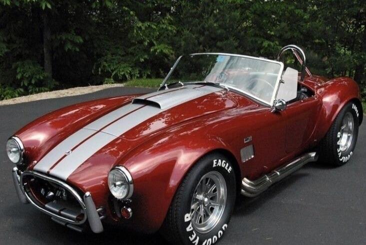 A classic red sports car with silver racing stripes, possibly a Shelby Cobra, parked outdoors. The tires have 'EAGLE' and 'GOOD YEAR' written on them.