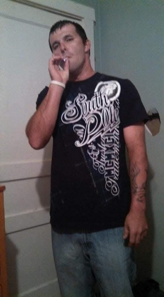 A man is standing, holding a cigarette or joint to his mouth. He is wearing a black t-shirt with 'South Pole' text and blue jeans. He has a white wristband and a tattoo on his arm.
