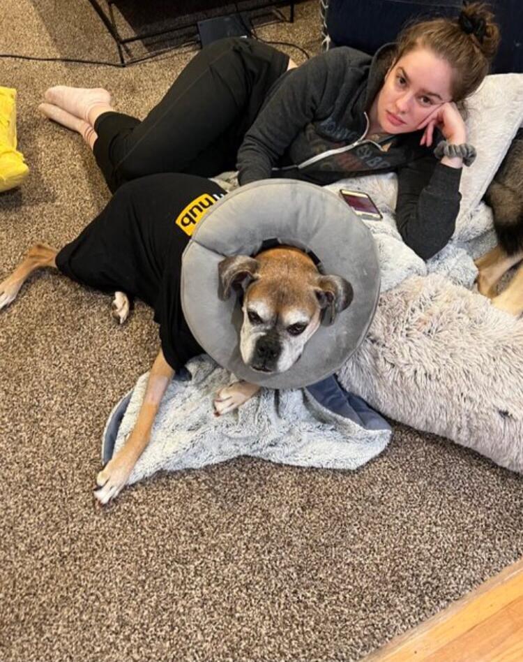 A woman is lying on the floor next to a dog wearing a cone of shame and a black t-shirt with 'hub' written on it.