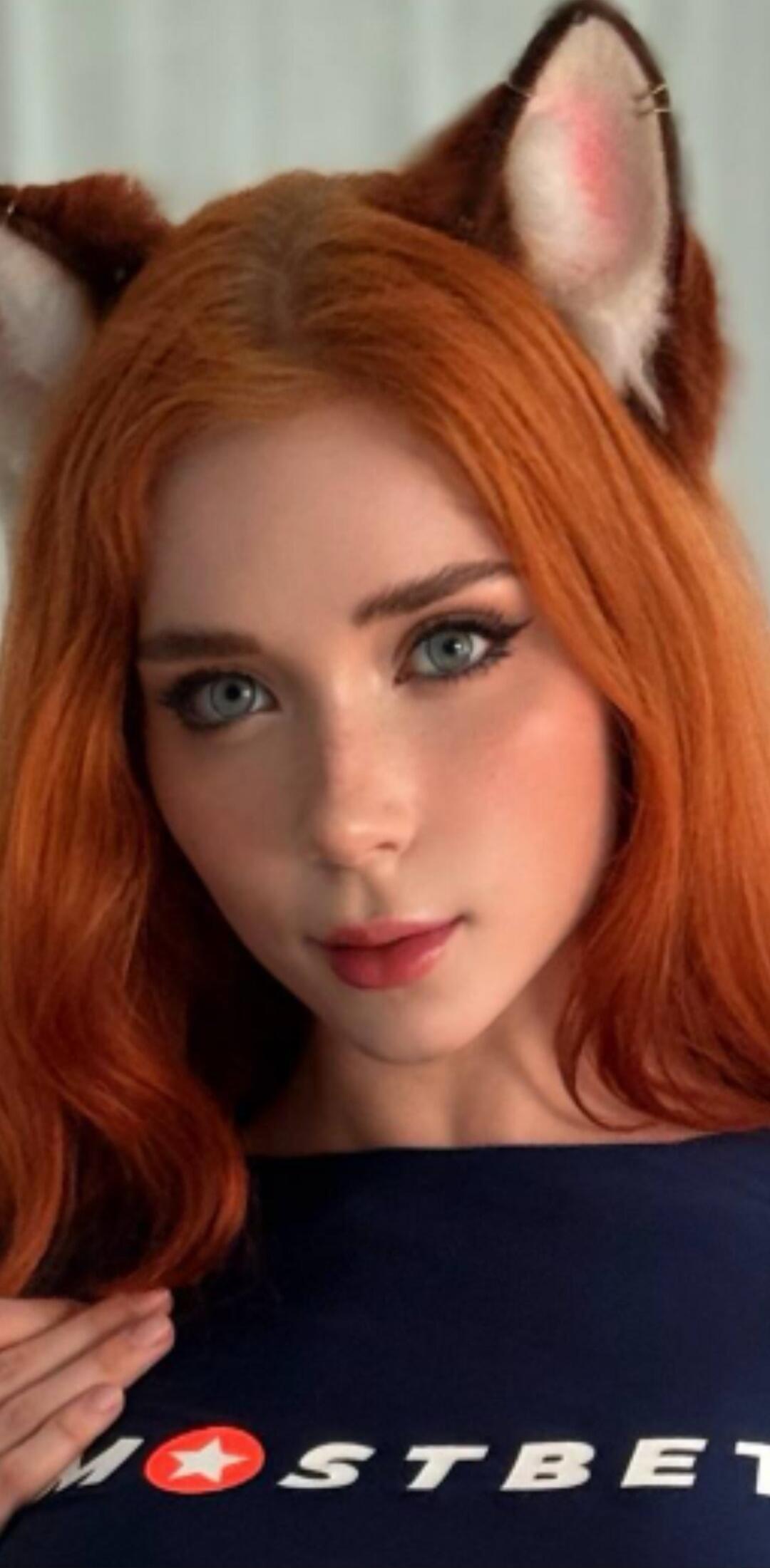 A young woman with red hair and blue eyes, wearing fox ears and a dark blue top with the 'MOSTBET' logo.