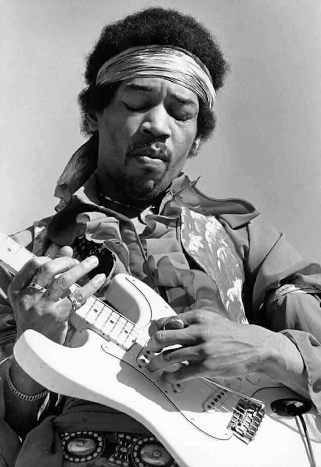A black and white photo of Jimi Hendrix playing an electric guitar with his eyes closed, wearing a headband and a ruffled shirt.