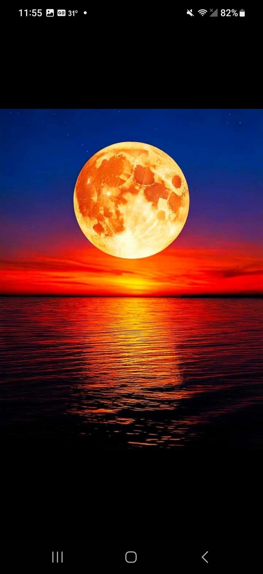 11:55 31° 82%. A large, glowing orange full moon hangs over a calm body of water, reflecting the vibrant red and orange hues of a sunset or sunrise sky. The sky transitions from deep blue at the top to fiery red and orange near the horizon.
