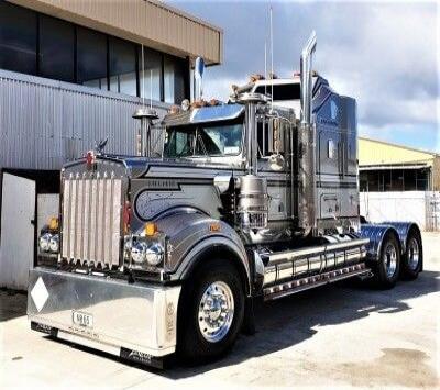KENWORTH W900 chrome tractor-trailer with a long-nose design, parked outside an industrial building. Prominent chrome details, dual rear axles, and polished front grille.