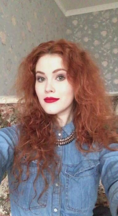 Photo of a woman with long, curly red hair wearing a blue denim shirt.