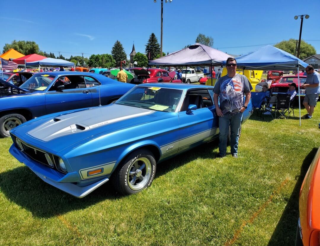 Blue classic muscle car on a sunny day at a car show, with a man standing beside it. Tents and other cars in the background.